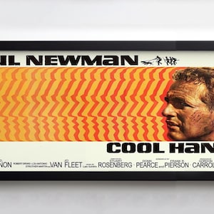 May include: Framed movie poster for "Cool Hand Luke" featuring Paul Newman. The poster has a black frame and a cream-colored background. The title and actor's name are in bold black text. The poster has a distinctive orange and yellow design.