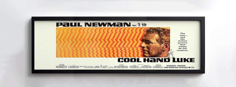 May include: Framed movie poster for "Cool Hand Luke" featuring Paul Newman. The poster has a black frame and a cream-colored background. The title and actor's name are in bold black text. The poster has a distinctive orange and yellow design.