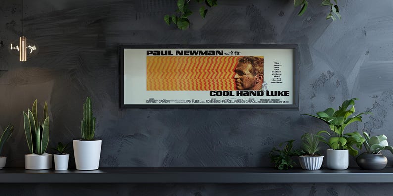 May include: A framed movie poster for "Cool Hand Luke" featuring Paul Newman. The poster has a black frame and is displayed on a dark gray wall. The poster's design includes orange and yellow stripes and a profile of Newman's face.
