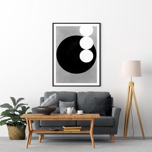 Op de afbeelding: Een ingelijste abstracte kunstprint met een groot zwarte cirkel en drie witte cirkels op een grijze achtergrond. Het kunstwerk hangt boven een grijze bank en past in een moderne woonkamer. De print zit in een zwarte lijst.