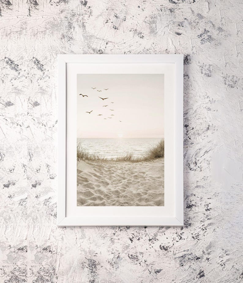 May include: A framed print featuring a serene beach scene. The image shows a sandy beach, calm ocean, and a flock of birds in flight against a soft, pastel sky. The print is encased in a white frame, creating a minimalist aesthetic.
