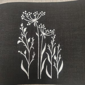 May include: Close-up of black fabric with a white floral pattern. The design showcases white flowers and leaves. The image highlights the fabric's texture and the detailed botanical illustration, creating a striking contrast.