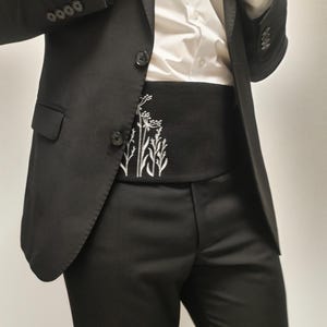 May include: A black suit with a white shirt and a black cummerbund. The cummerbund has a white embroidered floral design. The suit jacket has two buttons and the trousers are a classic fit.