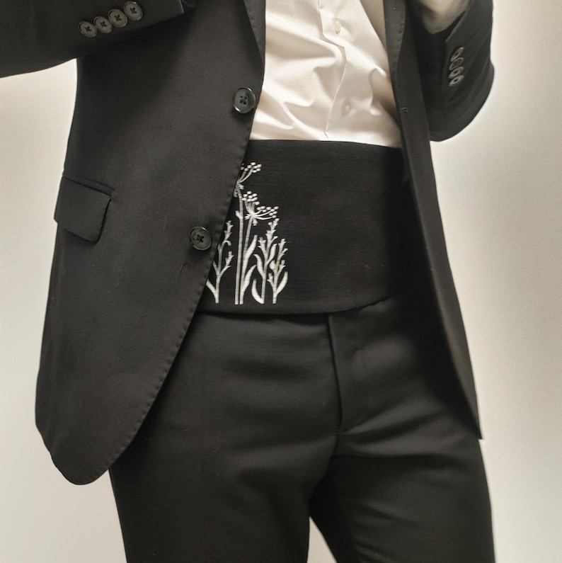 May include: A black suit with a white shirt and a black cummerbund. The cummerbund has a white embroidered floral design. The suit jacket has two buttons and the trousers are a classic fit.