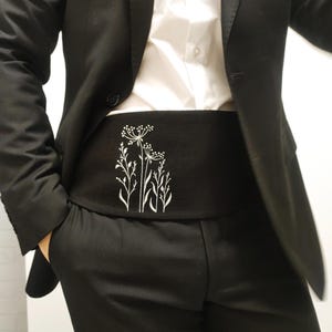 May include: Black cummerbund featuring a white floral pattern. The cummerbund is worn with a black suit and a white shirt. The floral design includes detailed stems and flower heads.