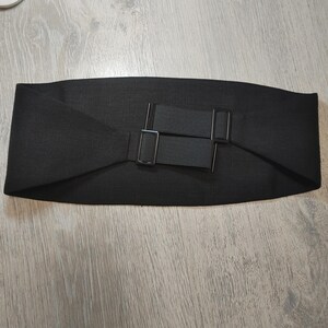 May include: A wide, black belt with a rectangular buckle and elastic straps. The belt is made of a textured fabric and is designed to cinch the waist. The buckle and straps are also black.