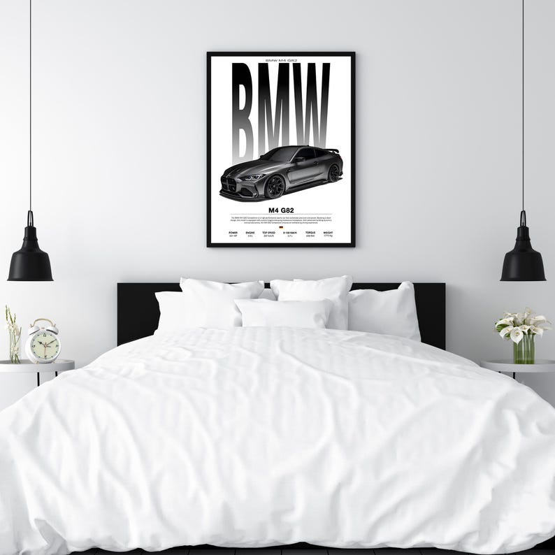 May include: A framed black and white poster featuring a BMW M4 G82 car. The BMW logo is prominently displayed above the car image. The poster is mounted on a white wall above a bed with white bedding.