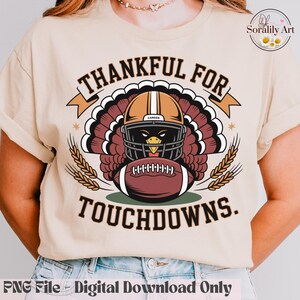 May include: Beige t-shirt with a Thanksgiving-themed graphic. The design features a turkey wearing a football helmet and holding a football, with the words "THANKFUL FOR TOUCHDOWNS." Wheat stalks and stars are also included in the design.