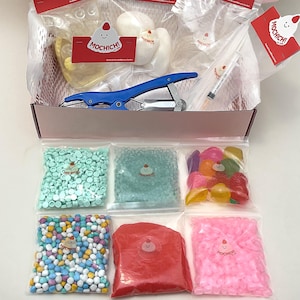 May include: A pink box filled with various craft supplies. The box contains bags of colorful beads, candies, and other materials. The box also includes a blue stapler, a syringe, and several small red cards with the text "Mochichi!" and a smiling face.