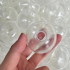Op de afbeelding: Een heldere, ronde glazen ornament met een gat in het midden wordt in een hand gehouden. De ornament wordt omringd door vele andere soortgelijke heldere glazen ornamenten. De ornamenten lijken voor knutselen of decoratie te zijn.