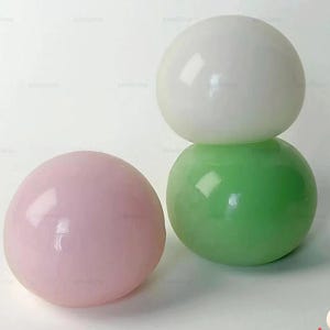 May include: Three colourful, spherical mochi desserts. One pink mochi is on the left, a green mochi in the middle, and a white mochi stacked on top of the green one. The mochi are smooth and glossy. The image includes the text "MOCHICH!" by dotta.