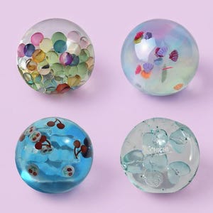 May include: Four clear spherical balls with embedded decorations. The top left ball contains multicolored circular confetti. The top right ball features small colorful seashells. The bottom left ball has red cherry motifs. The bottom right ball displays light blue shapes and the text OCHICHI.