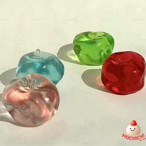 Peut inclure: Quatre objets en forme de pomme translucides et colorés, dans les tons rose, bleu, vert et rouge. Chacun a une petite tige et projette une ombre. Les objets sont disposés sur une surface claire.