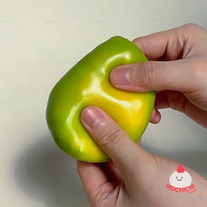 May include: A smooth, pliable, lime green and yellow slime toy with a glossy finish. The material is being pressed and stretched, showing its flexible texture. In the bottom right corner, there is a small logo with the text MOCHICH! by Joli.