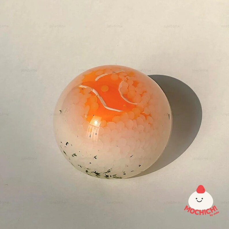 May include: A clear, spherical object with an orange interior design and white accents. The bottom portion has small green and black specks. A shadow is cast to the right. The image has a clean, minimalist aesthetic.