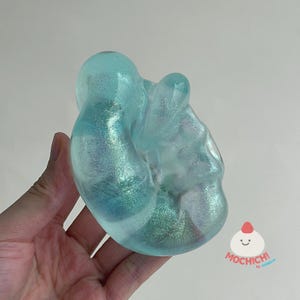 May include: A translucent, light blue, amorphous resin object with a shimmering, glittery finish throughout. The surface is smooth and rounded with organic, irregular curves. A small logo with a smiling face and the text MOCHICH! is visible.