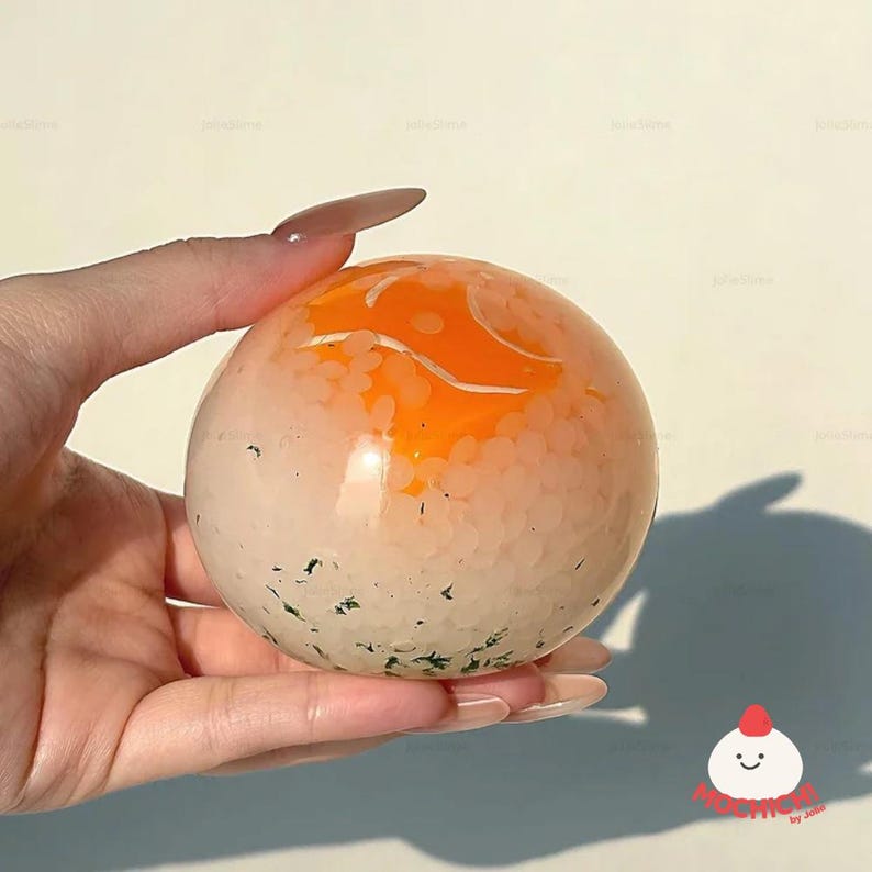 May include: A spherical, translucent object with an orange top and white base, speckled with green and white details. The object is held in a hand, showcasing its smooth, rounded form. The object has a unique design.
