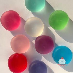 May include: A collection of nine smooth, round, translucent silicone squishy toys in various colors including red, pink, light blue, dark blue, purple, green, teal, and white. A small logo with a cartoon character and the text MOCHICHI is visible.
