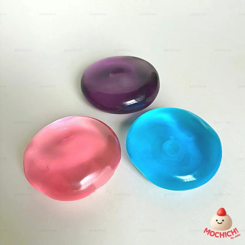 May include: Three smooth, colorful, gel-like stones in shades of pink, blue, and purple. The stones have a translucent appearance and a rounded, organic shape. The image includes a logo in the bottom right corner.