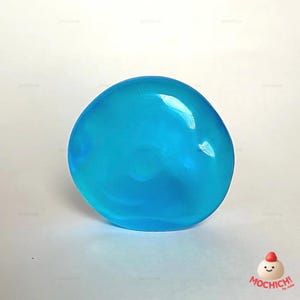 Op de afbeelding: Een helder, doorschijnend, blauw, bolvormig object. Het object heeft een glad, glanzend oppervlak en lijkt een gel of een vergelijkbare substantie te zijn. Het object staat tegen een effen witte achtergrond. De afbeelding bevat de tekst "MOCHICH!" en een klein stripfiguur.