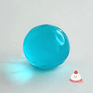 Op de afbeelding: Een heldere, ronde, blauwe gelbal met een glanzend oppervlak. De gelbal werpt een schaduw op een lichte achtergrond. De afbeelding bevat de tekst "MOCHICH! by Jolie" in de rechter benedenhoek.