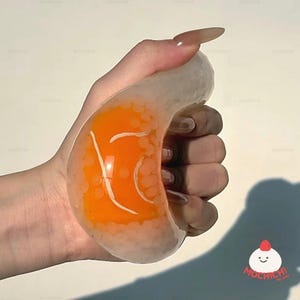 May include: A clear, curved stress ball filled with orange liquid and white beads. The orange liquid has white line designs. The stress ball is held in a hand. The logo "Mochichi" is in the bottom right corner.