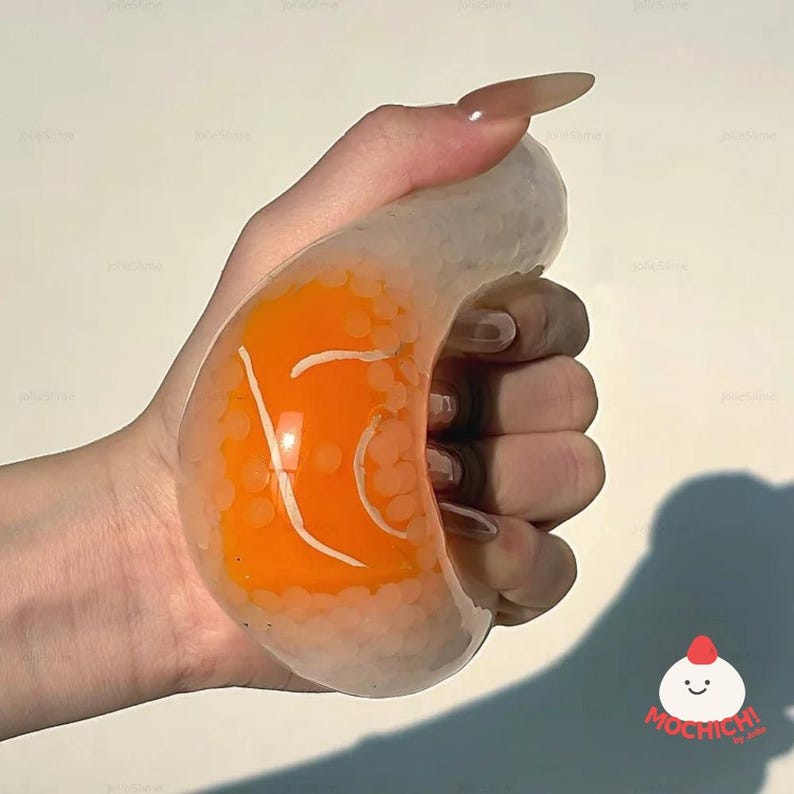 May include: A clear, curved stress ball filled with orange liquid and white beads. The orange liquid has white line designs. The stress ball is held in a hand. The logo "Mochichi" is in the bottom right corner.