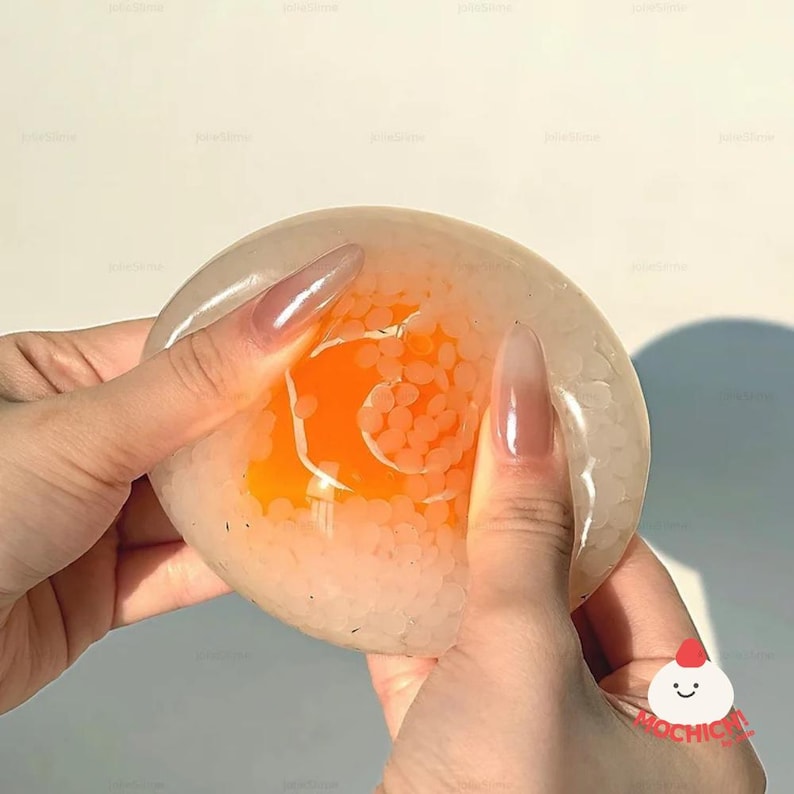 May include: A clear, round stress ball filled with orange and white beads. The ball is being held by two hands with long, polished nails. The stress ball has a bright orange center.