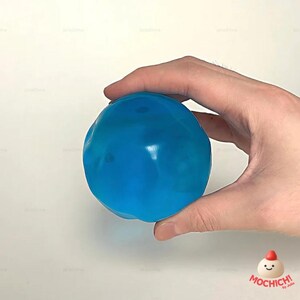 Op de afbeelding: Een helderblauwe stressbal met een glad, glanzend oppervlak, vastgehouden in een hand. De bal heeft een onregelmatige vorm. De achtergrond is effen wit, wat de kleur en vorm van de bal benadrukt.