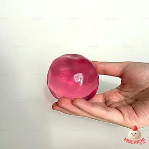 Op de afbeelding: Een hand houdt een doorschijnend, roze, bolvormig object vast tegen een witte achtergrond. Het object lijkt op gel of slijm. Een logo is zichtbaar in de rechter benedenhoek van de afbeelding.