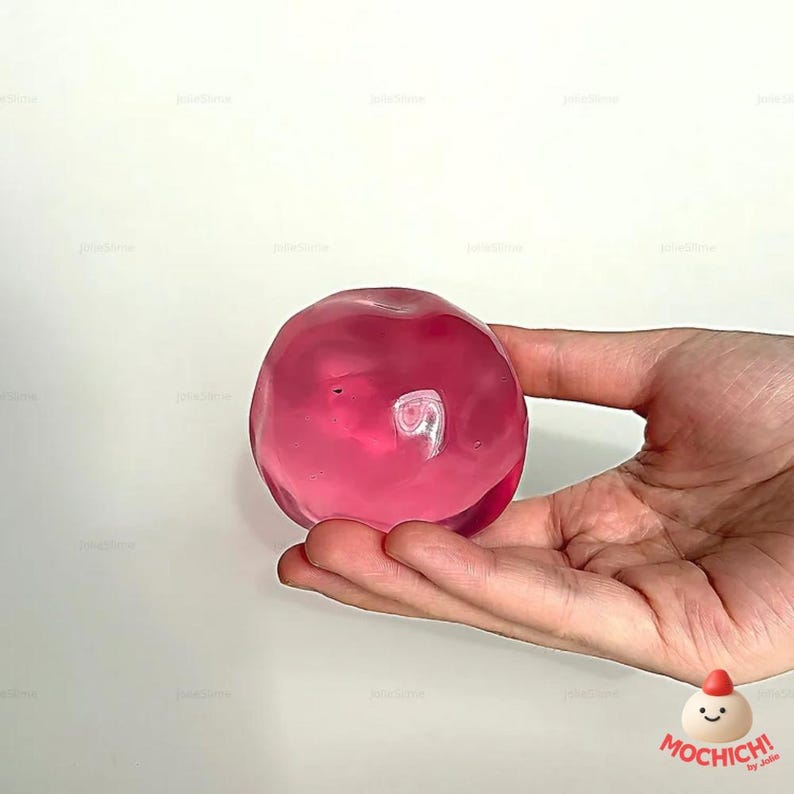 Op de afbeelding: Een hand houdt een doorschijnend, roze, bolvormig object vast tegen een witte achtergrond. Het object lijkt op gel of slijm. Een logo is zichtbaar in de rechter benedenhoek van de afbeelding.