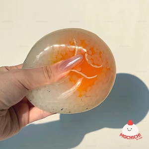May include: A clear, squishy slime toy with white beads and an orange center. The slime is held in a hand, showing its texture and color. The toy has a logo in the bottom right corner.