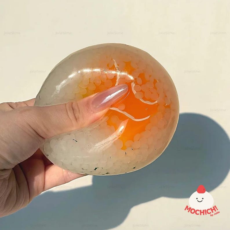 May include: A clear, squishy slime toy with white beads and an orange center. The slime is held in a hand, showing its texture and color. The toy has a logo in the bottom right corner.