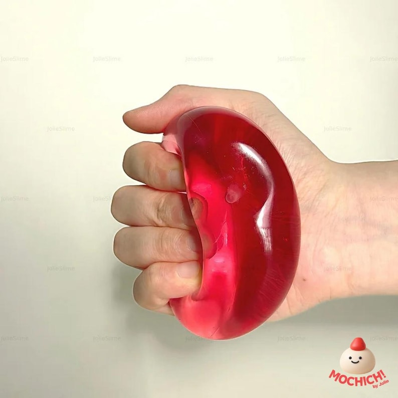 Op de afbeelding: Een hand knijpt in een doorschijnende, rode, boonvormige stressbal. De stressbal is glad en glanzend, met een lichte kromming. De achtergrond is effen, gebroken wit. Het logo "Mochich!" staat rechtsonder.