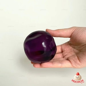 Op de afbeelding: Een dieppaars, bolvormig object met een glad, glanzend oppervlak. Het wordt in een hand gehouden tegen een witte achtergrond. Het object lijkt een decoratief item of een speelgoed te zijn. Ongeveer 7 cm in diameter.