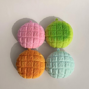 Puede incluir: Cuatro juguetes redondos y texturizados dispuestos en formación cuadrada. Cada artículo presenta un patrón de cuadrícula en su superficie y un pequeño lazo en la parte superior. Los colores incluyen rosa pastel, verde lima vibrante, naranja cálido y azul claro.