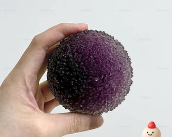 May include: A textured, spherical purple stress ball. The ball is held in a hand against a white background. The ball has a rough, bumpy surface. The logo "Mochich! by Jolie" is in the bottom right corner.