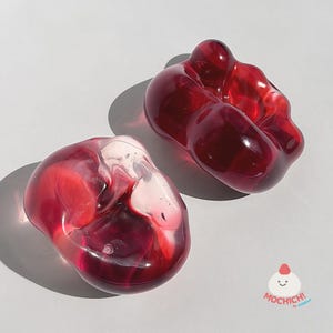 May include: Two translucent, organic-shaped objects made of red glass. One piece is a deep, solid crimson, while the other features a swirling blend of clear and dark red tones. Both items have smooth, rounded surfaces and a glossy, reflective finish.