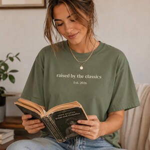 May include: An olive green t-shirt with the text "raised by the classics" and "Est. 2026" printed on the front. The person is wearing the shirt and reading a copy of "Pride and Prejudice" by Jane Austen.