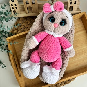 Può includere: Bambola coniglietto all'uncinetto fatta a mano con corpo rosa, dettagli bianchi e fiocco rosa. Il coniglietto ha grandi occhi blu ed è appoggiato su un vassoio di legno. Case decorative in legno sullo sfondo.