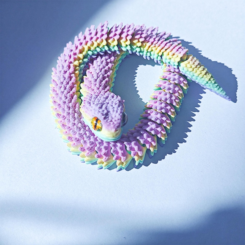 May include: A colorful, articulated snake toy in shades of purple, yellow, and blue. The snake is coiled, with a textured body and a detailed head featuring an orange eye. The toy is designed to be flexible and poseable.