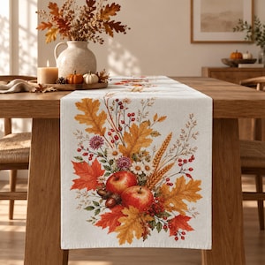 Könnte beinhalten: Ein cremefarbener Tischläufer aus Leinen mit einem herbstlichen Druck aus roten Äpfeln, goldenen Eichenblättern, Ahornblättern, Weizenähren, kleinen weißen Blumen, violetten Blüten und roten Beeren in einem dekorativen Erntemotiv.