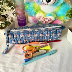 May include: A blue floral pencil case with a zipper, next to a collection of pens, highlighters, and scissors. A colorful plush bear and a water bottle with text are in the background. The items are arranged on a white surface.
