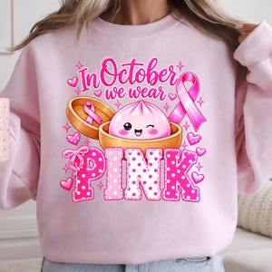 In October We Wear Pink breast cancer awareness PNG featuring a cute pink dumpling in a bamboo steamer, pink ribbon, hearts, bows, sparkles, and polka-dot lettering.