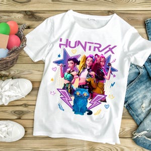 A product mockup showing a vibrant Huntrix Kpop Demon Hunters graphic applied to a physical item. The print highlights the vivid colors and crisp details of the Saja Girls anime PNG design, demonstrating its perfect application for sublimation and DTG printing.