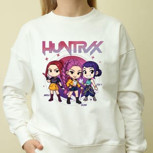 A product mockup showing a vibrant Huntrix Kpop Demon Hunters graphic applied to a physical item. The print highlights the vivid colors and crisp details of the Saja Girls anime PNG design, demonstrating its perfect application for sublimation and DTG printing.