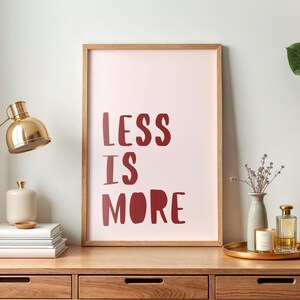 Könnte beinhalten: Ein Holzschreibtisch mit einem gerahmten Druck mit der Aufschrift "LESS IS MORE" in burgunderroten Buchstaben. Eine goldene Schreibtischlampe, Bücher und dekorative Gegenstände stehen auf dem Schreibtisch. Eine Pflanze ist rechts zu sehen.