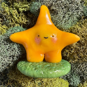 May include: A smooth, bright orange star-shaped figurine with a glossy finish, featuring two small black eyes, a simple curved black mouth, and soft pink blush on its cheeks. The star stands upright on a textured, round, light green base.