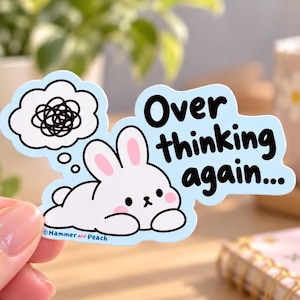May include: A light blue die-cut sticker featuring a white cartoon rabbit lying down with pink cheeks. Above the rabbit is a thought bubble containing a black scribble. The text reads Over thinking again... with Hammer and Peach printed below.