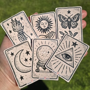 May include: A collection of six rectangular stickers with a light beige background and black ink illustrations. Designs include a hand holding a torch, a sun, a moth, a crescent moon, a crystal ball, and an eye within a triangle. Each sticker features the text Hammer Peach.